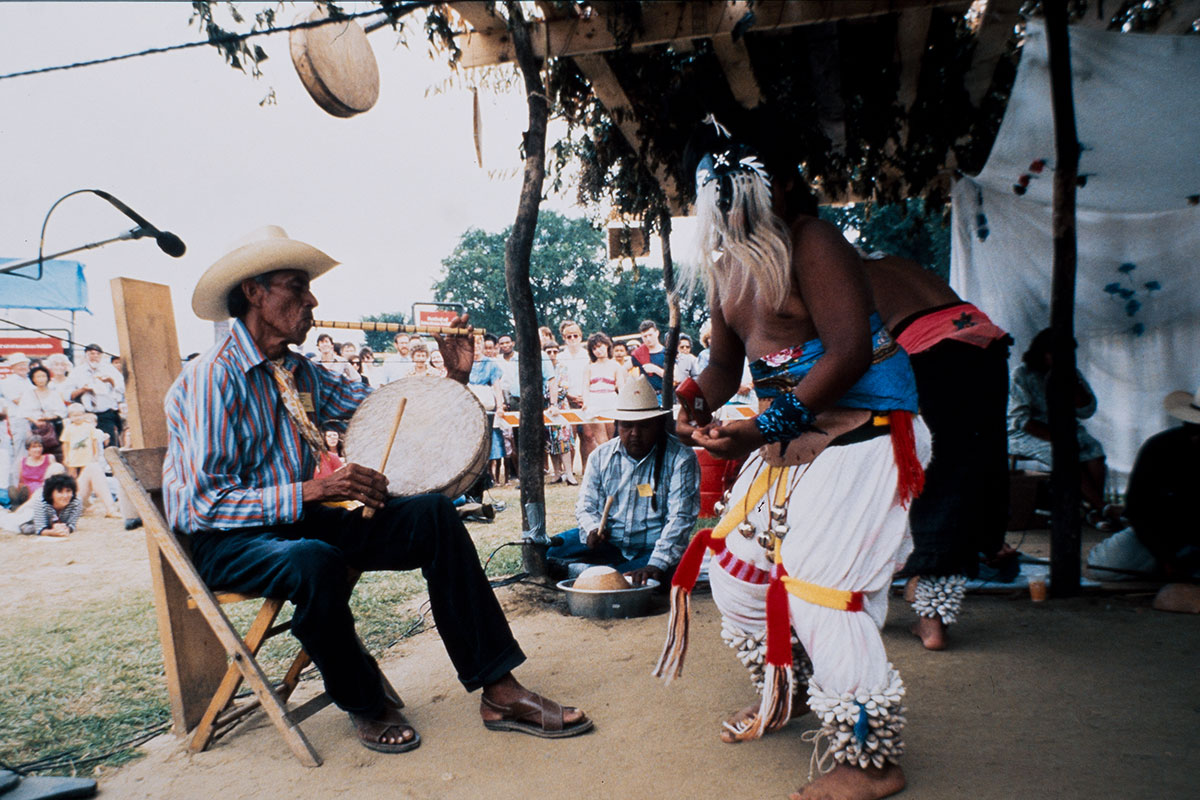 Three performers on an outdoor stage: a man seated in a chair playing a frame drum and a whistle; a person dancing with jingle anklets, and a person seated on the ground playing a gourd drum.