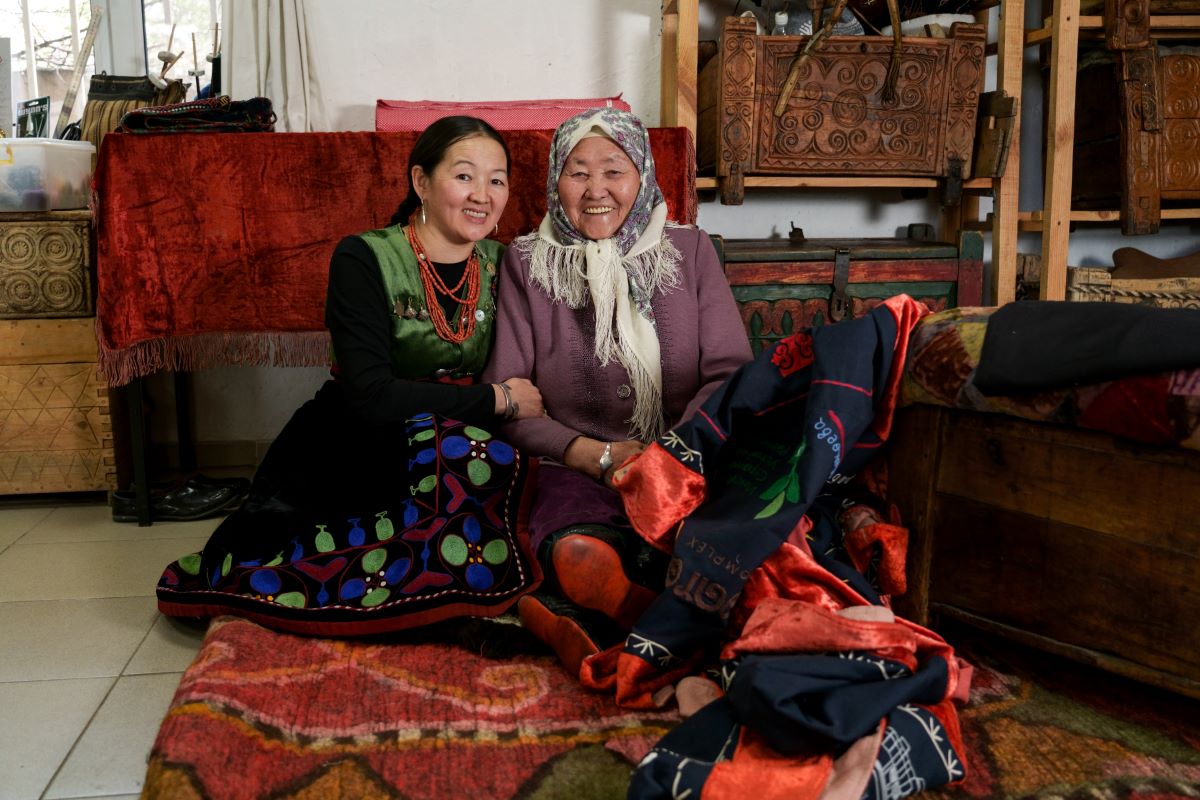 Two adults sit on the floor while holding each other and smiling at the camera. They are dressed in traditional outerwear from Kyrgyzstan.