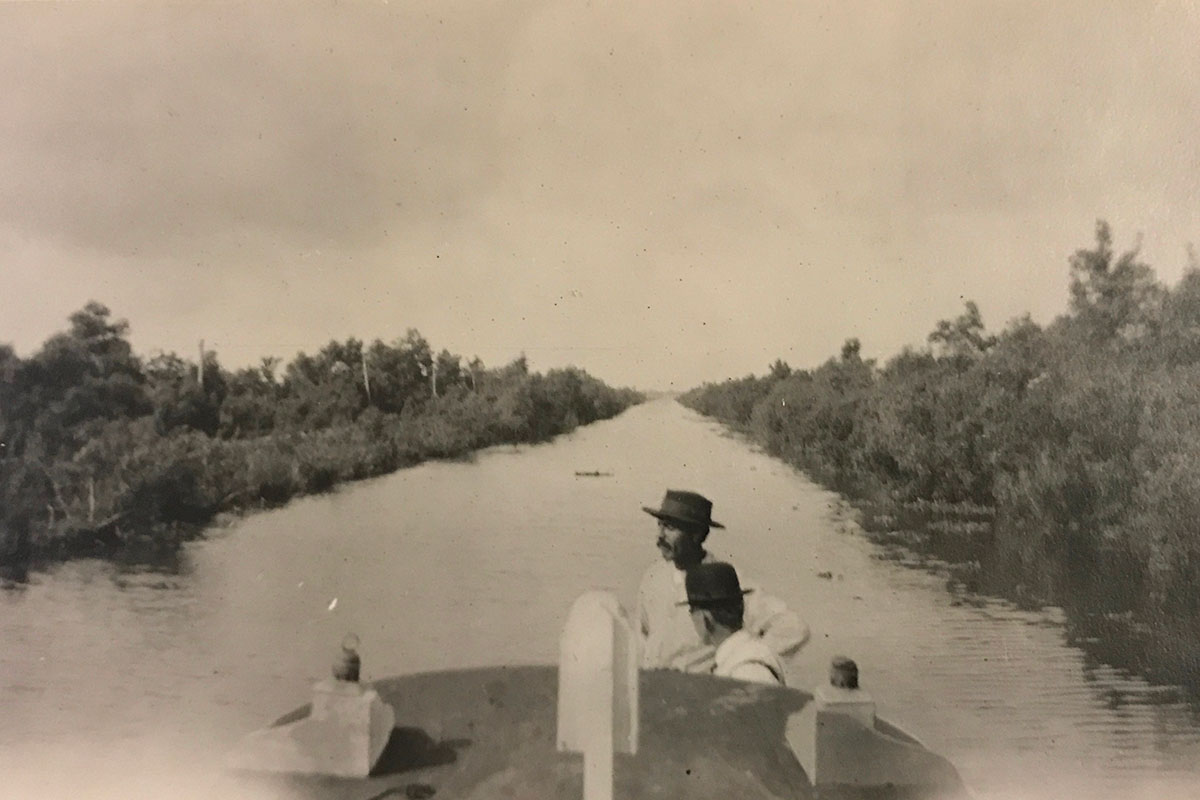 Two people on a boat in the middle of a tree-lined river. Black-and-white photo.