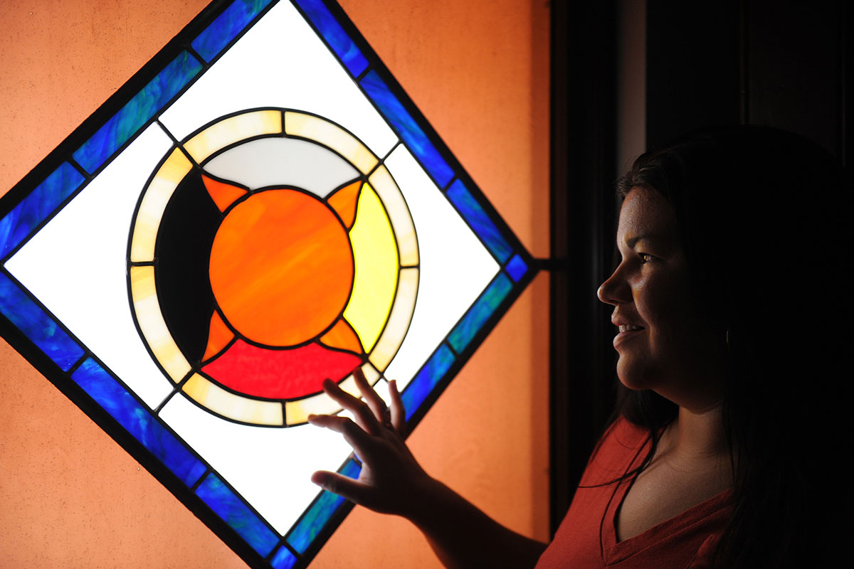 A woman's face is illuminated by the light coming through a stained glass window. Its pattern shows an orange sun with four points, and each quarter section in a different color: light blue, yellow, red, and black. 