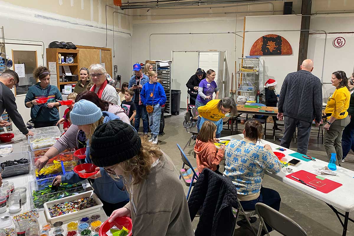 Kids and adults work on various tables in a workshop room. One table is lined with bins of colored glass pieces.