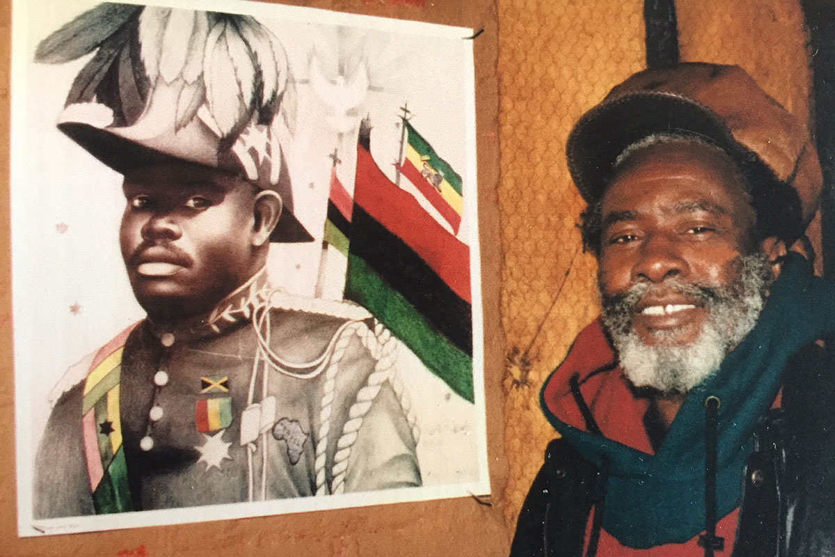 A Black man smiles, posing next to a poster of a Black man in military dress and the Pan-African flag.
