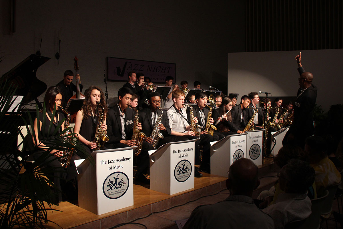 More than twenty teen perform on stage, most in black dress. Their podiums read The Jazz Academy of Music.