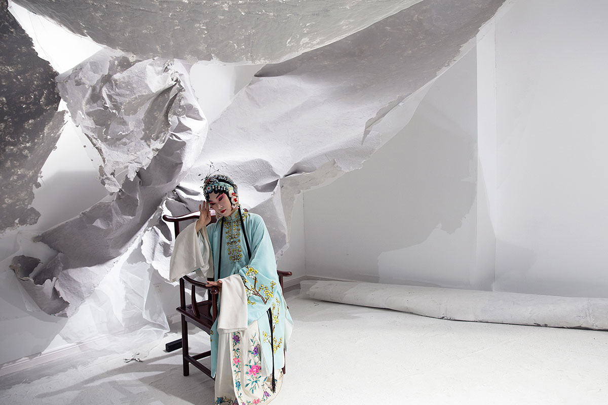 An Asian woman wearing exaggerated stage makeup and a sky-blue embroidered robe sits in a chair, contrasting with a white sculptural piece of hung, torn, and crumpled paper or fabric.