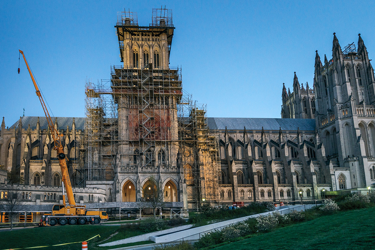 Against a dusky dark blue sky, the National Cathedral stands blocked by extensive scaffolding and a tall yellow crane. 