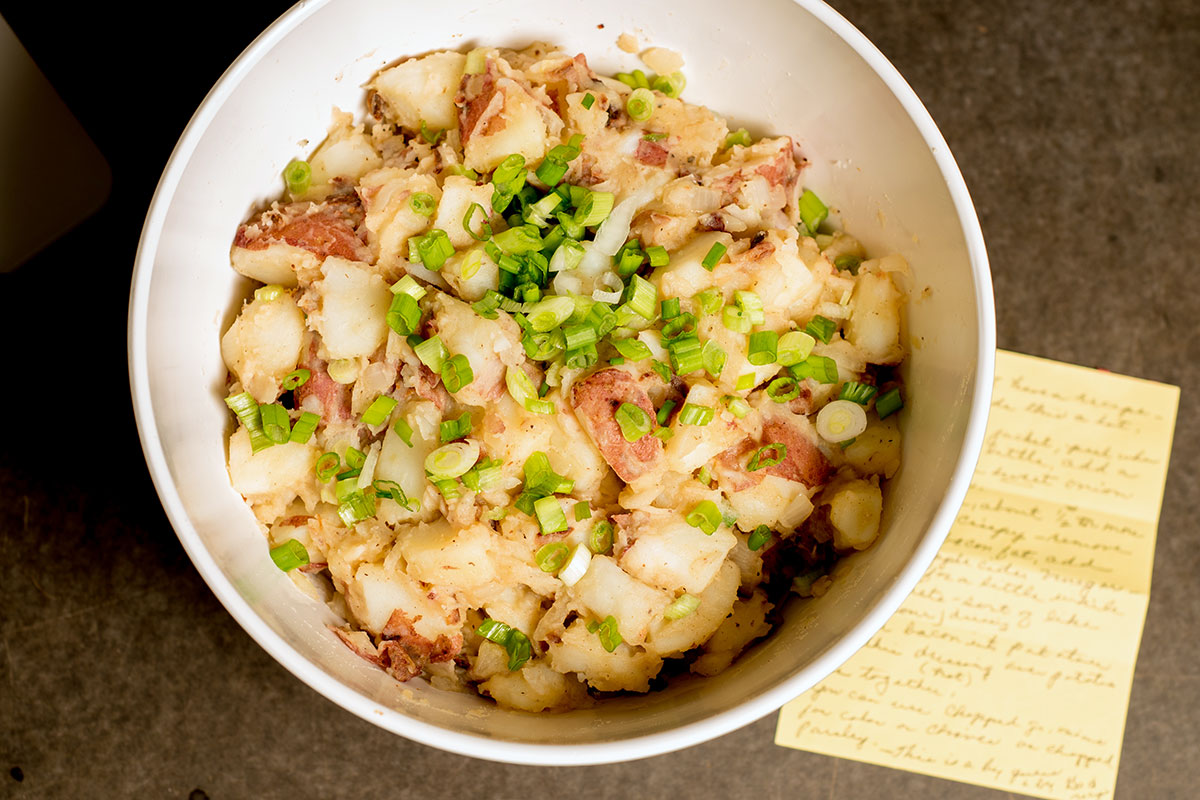 Bowl of potato salad topped with chopped green onion, alongside a handwritten recipe on a notecard.