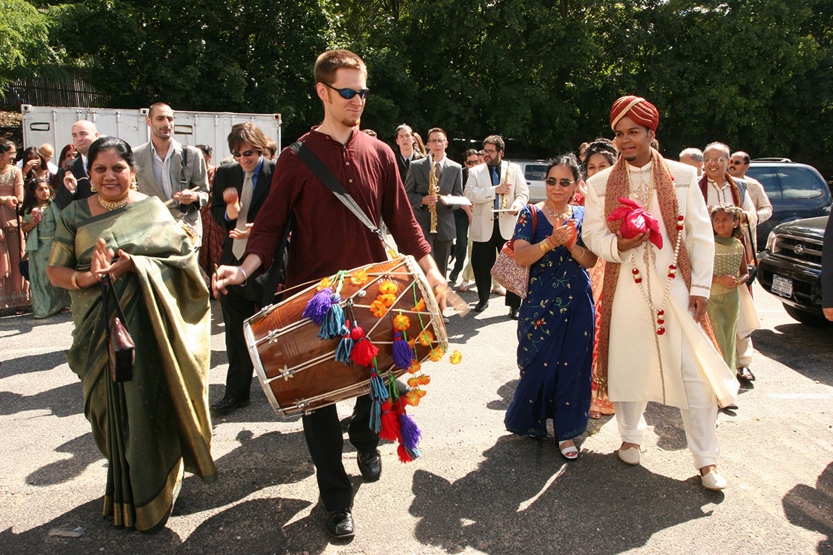 The front of a wedding procession through a parking lot, led by a white man in the center playing a double-headed barrel-shaped drum adorned with orange flowers and multicolored tassels of yarn. To the right is the groom (the author) in traditional Punjabi wedding clothes: white tunic, pants, and shoes, a red and gold headdress, and white and gold jewelry. The crowd following them is a mix of South Asian and white people. Some are carrying musical instruments.