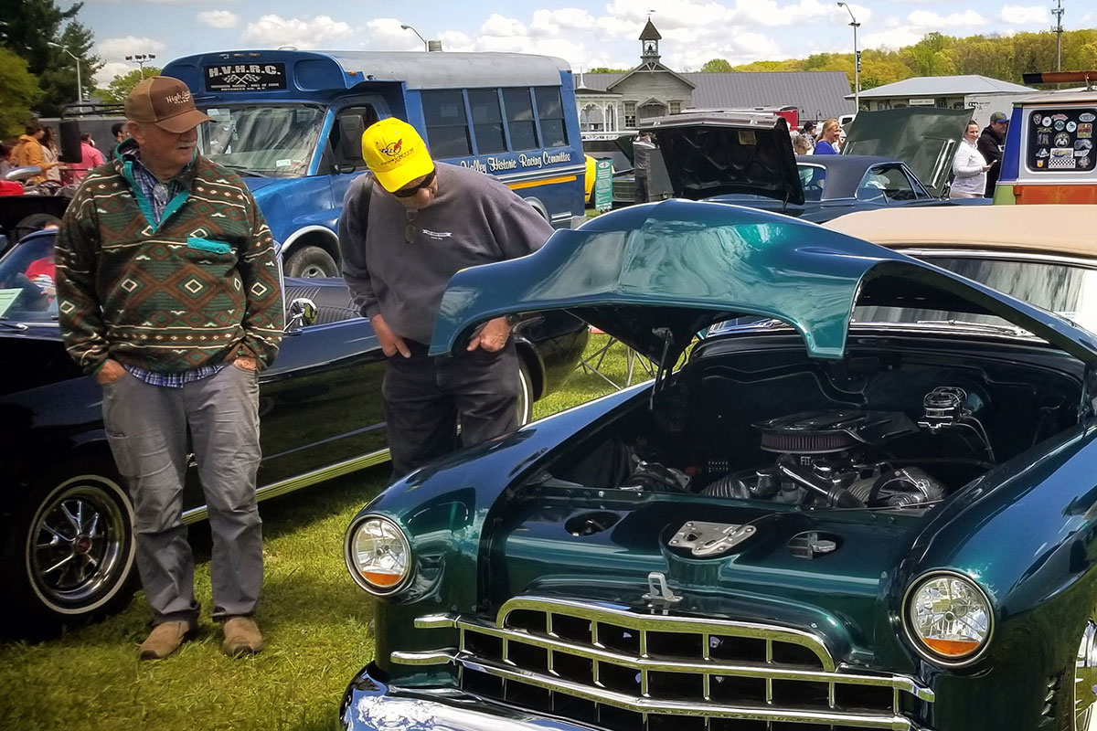 Two men inspect under the hood of a dark teal classic car, parked on a green lawn with other old vehicles.