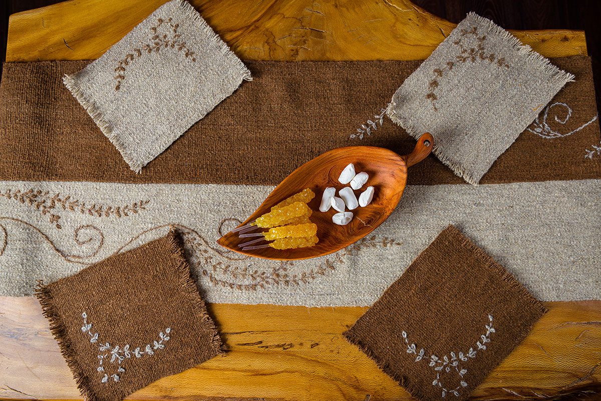 From overhead, a wooden table set with a wool runner in brown and cream and four  matching wool coasters, all embroidered with a vine pattern. In the center of the table, a wooden leaf-shaped dish holds yellow rock candy and white tablets.