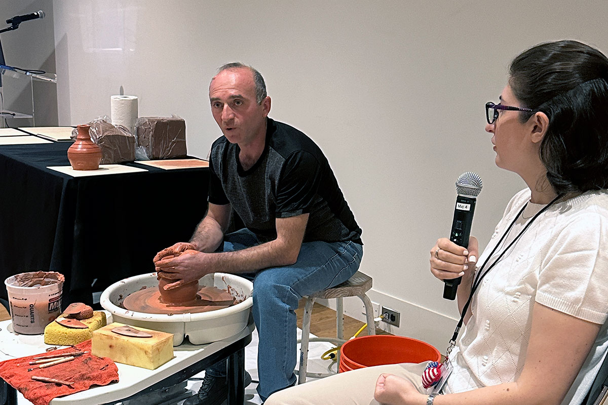 A man sits at a pottery wheel working red clay while a woman seated next to him speaks into a microphone.