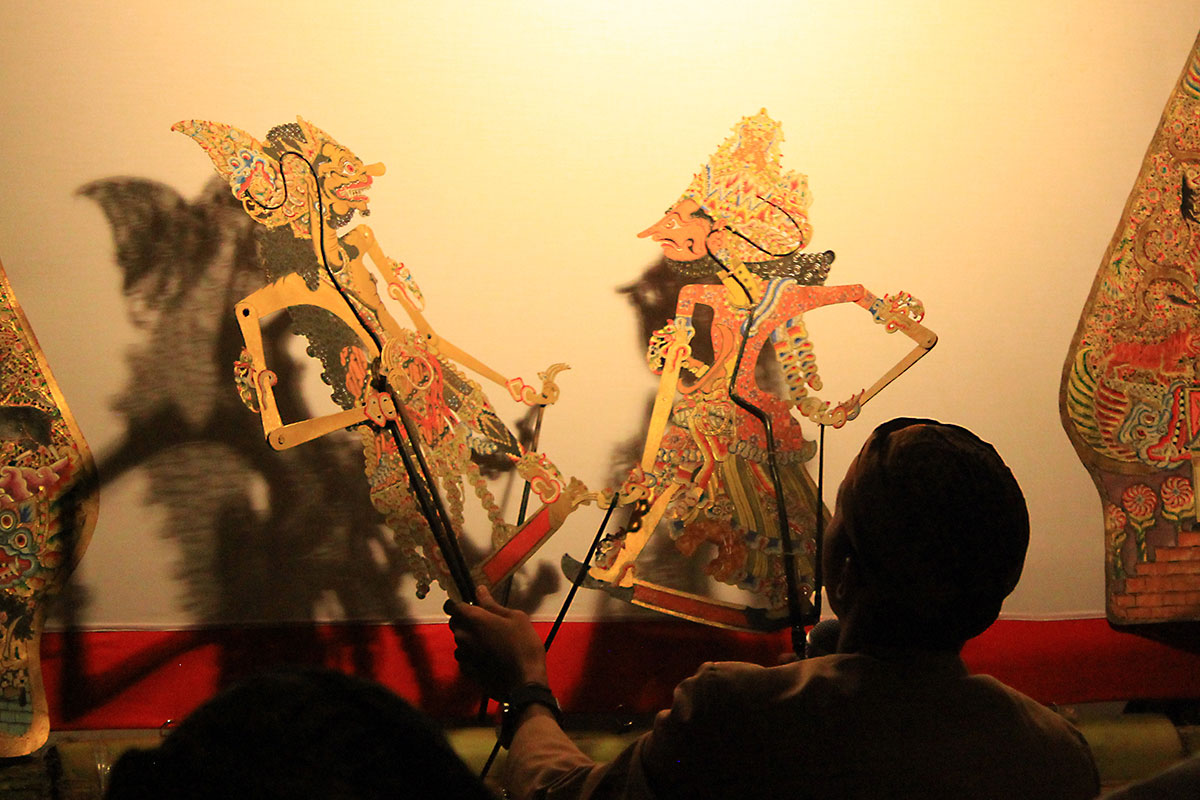 A person seated, facing away, maneuvers two intricately carved and colored two-dimension shadow puppets against a white screen. 