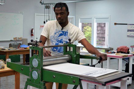 A young man pulls a crank on a green printing press in a studio work space. He wears headphones which rest on the collar of his shirt before going up to his ears.