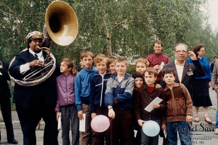A man dressed in a black suit blows into a sousaphone, posing with a group of young kids.
