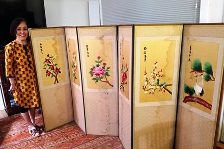 A woman in yellow and black patterned dress stands to the left of a roughly five-foot-tall folding privacy screen. Each panel has a different colored flower embroidered on it, and the panels show water damage.