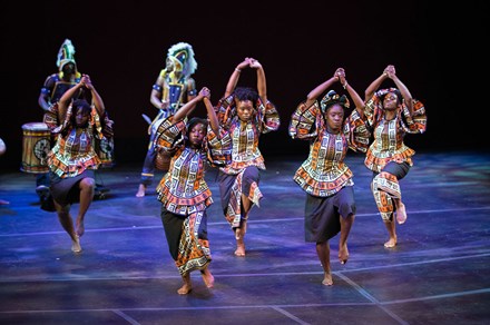Seven African American people dance on a stage, hands clasped overhead, wearing traditional printed blouses and skirts. Black backdrop.