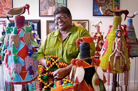 An African American woman poses and smiles among colorful artwork: paintings in the background, fabric sculptures in the foreground, and she is holding an African patterned textile.