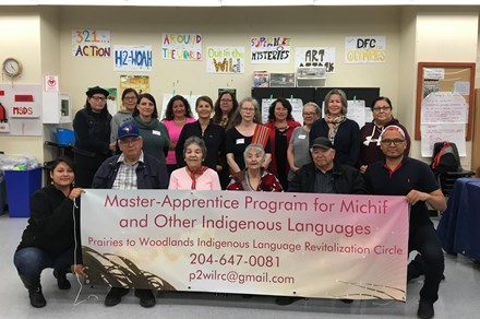 A multigenerational group of seventeen people stand in three rows for a photo. They are indoors, in a classroom and are holding a sign that reads: Master-Apprentice program for Michif and Other Indigenous Laguages.