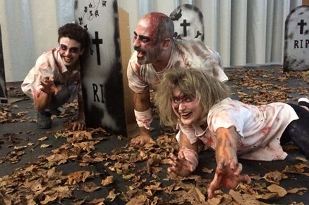 Three people act as blood-splattered zombies in a street filled with dead leaves and cardboard gravestones. 