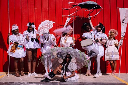 Eight people, both women and men, pose against a red wall, each dressed in a frilly pale pink dress with bonnets or large bows on their heads. Some carry decorative parasols. Some carry small dolls. 