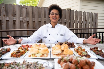 A Black woman in a white chef's coat opens her arms toward a table full of food.