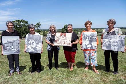 Six elder Black women pose on a grassy hillside, each holding an enlarged black-and-white photo of a schoolhouse, protestors with signs, and a class photo.