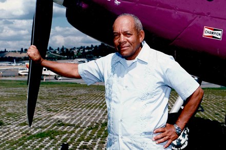 An older James Wiley poses, leaning on the propeller of a purple airplane, airfield in the background.
