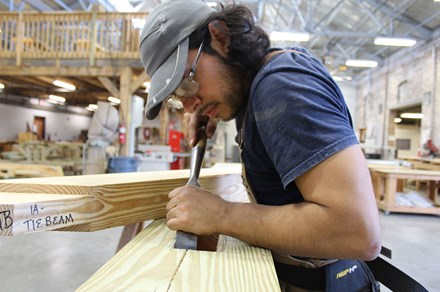 A young man fine tunes a cut in a piece of lumber using a hand tool that looks like a wide chisel.