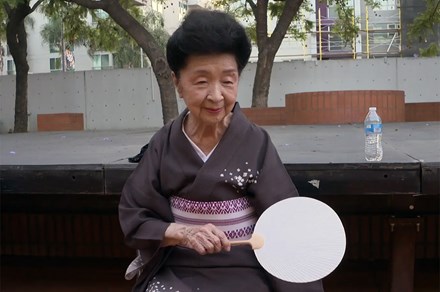 Madame Kansuma sits on an outdoor stage wearing a purple kimono, holding a white fan