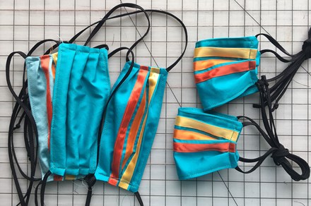 A stack of face masks on a gridded work station. They are turquoise with black straps and shiny ribbons sewn from side to side in stripes: red, orange, yellow.