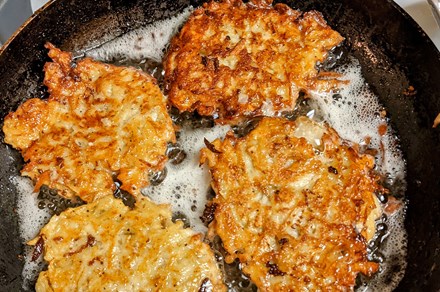 Four golden brown, shredded potato pancakes frying in a cast iron pan.