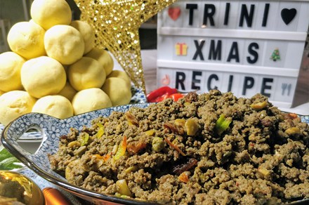 A bowl full of cooked ground meat, flecked with colorful vegetables pieces. In the background, a mound of yellow dough balls and a mini marquee-style sign that reads TRINI XMAS RECIPE.