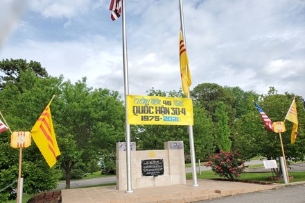 A beige stone memorial with a black plaque fixed on the front. In front of it, two flag poles, one with the American flag, and one with the South Vietnamese flag, yellow with three red stripes. A yellow banner between the poles has words in Vietnamese language and 1975-2021.