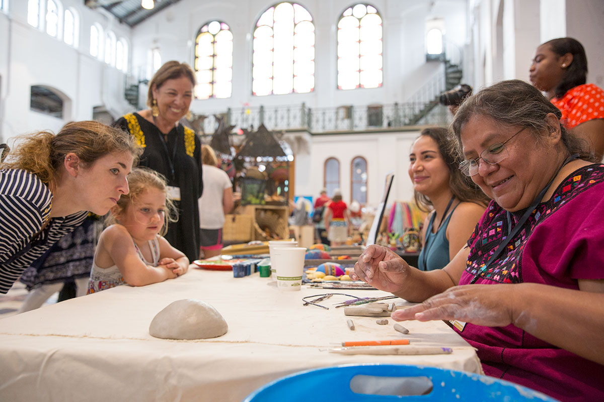 Women and children sit around a table indoors with a mound of clay and etching tools. 