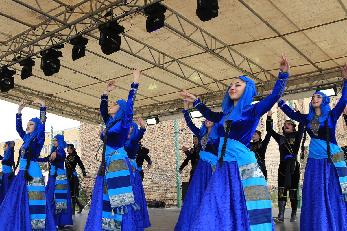 Several adults dance on a stage in loose blue outer garments. One individual stands in front of them in a black outfit, and several stand behind the group in similar dark outfits. They are dancing in unison and have their arms lifted in the air.