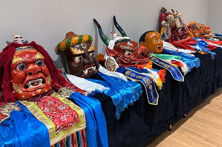 Display of six fanciful, intricately decorated carved masks on a table. Most are humanoid faces, plus one with a pig's features and two horns, and another of a furry animal. Some have three eyes.