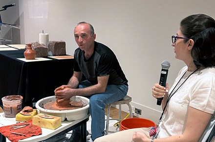 A man sits at a pottery wheel working red clay while a woman seated next to him speaks into a microphone.