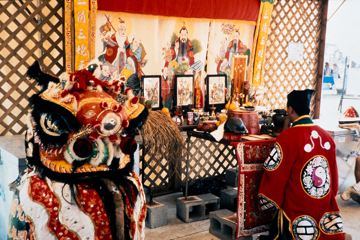 A man kneels at a table covered with incense, candles, and religious imagery, with a lion dance costume to his left.