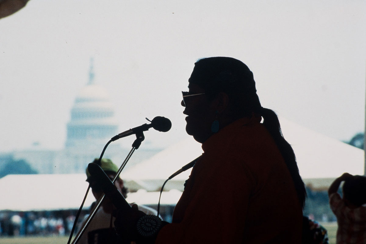 A silhouette of a man with a ponytail and aviator glasses faces left, against the backdrop of the U.S. Capitol Building. He leans into a microphone and holds a hand drum.