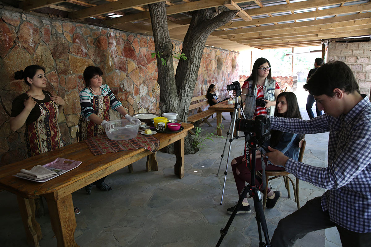 Two young women and a young man sit with video cameras and tripods filming two women wearing traditional Armenia dress on the right.