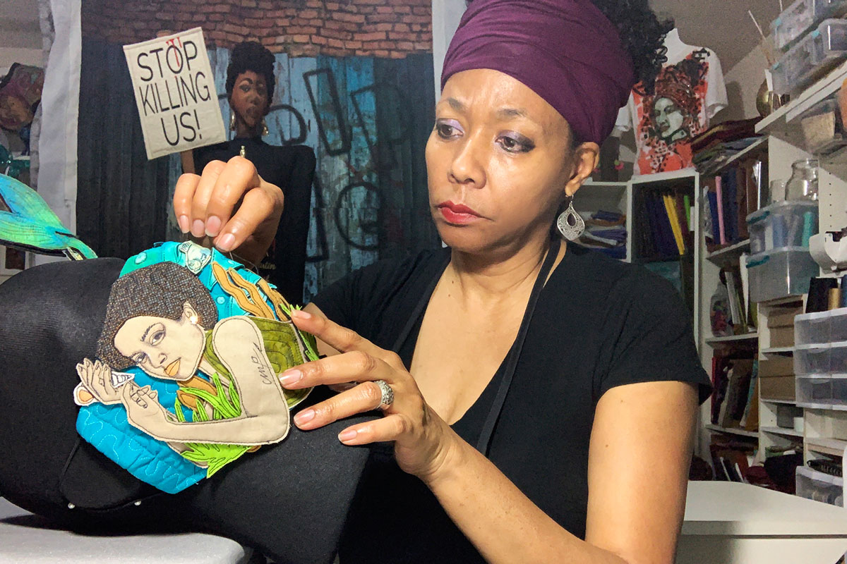 Artist Carolyn Crump sits and sews a face mask made of collaged fabric depicting a woman.
