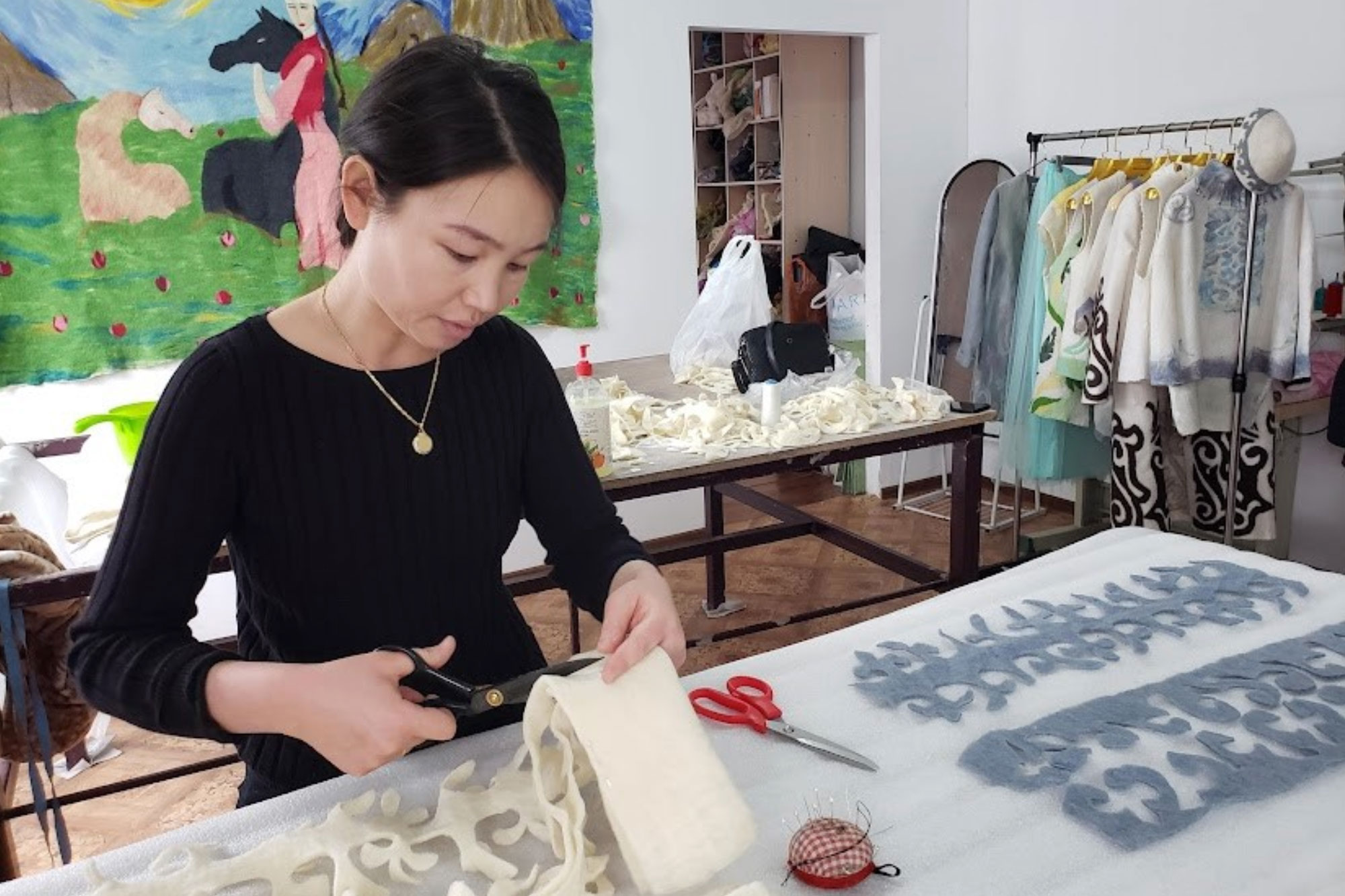 A woman in a brightly lit room stands behind a table and carefully cuts a cream-colored piece of felt. Beside her on both sides are long pieces of felt cut into vegetal motifs.