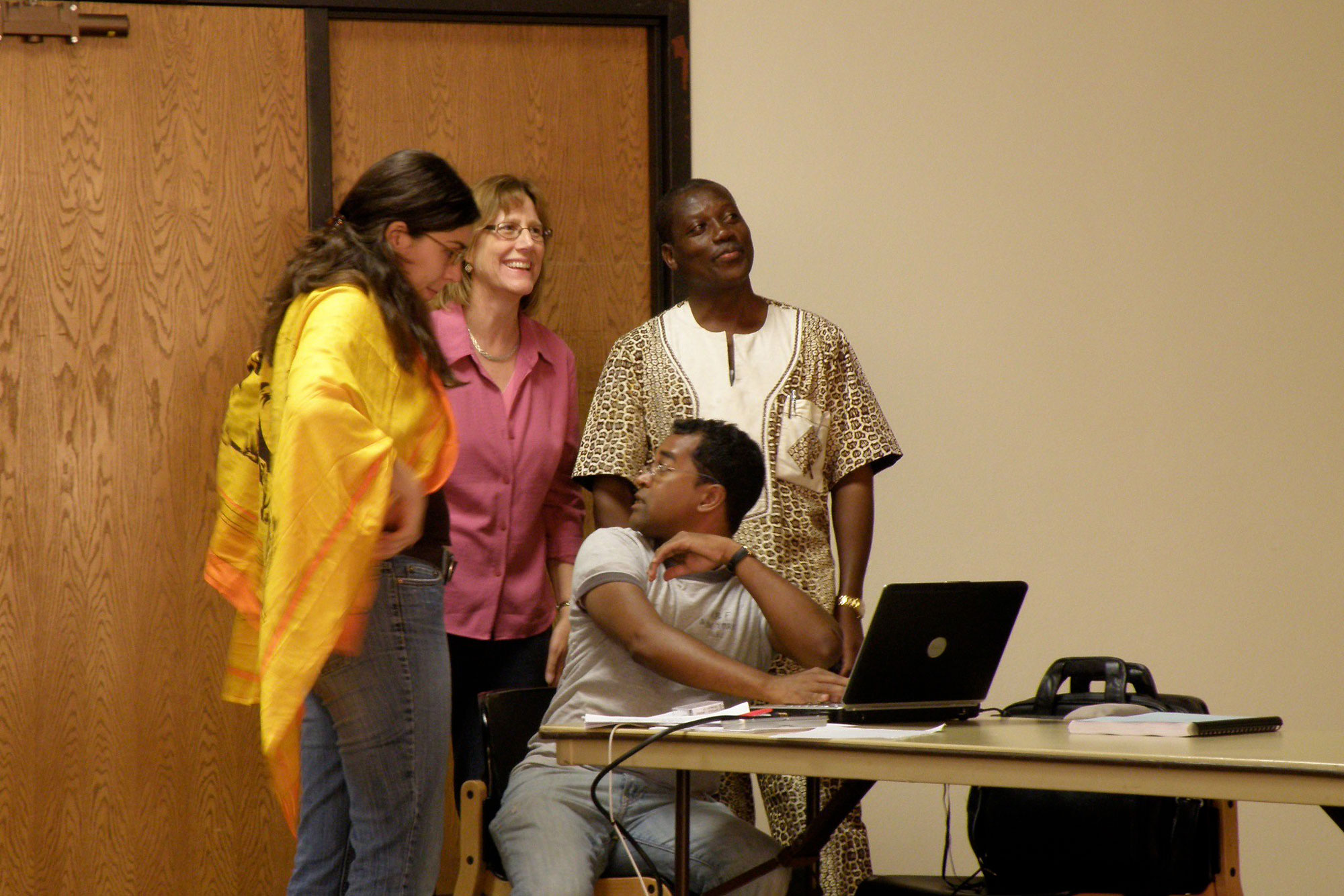 Two women and one man stand above a man seated at a desk with an open laptop. They look excited and in conversation as they ready to begin a presentation.