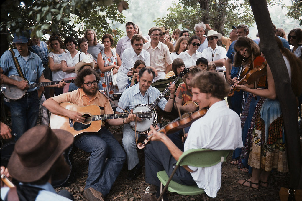 A group of people play music, with an acoustic guitar, banjos, and fiddles, in a circle outdoors under trees. A crowd of people gathers around them.