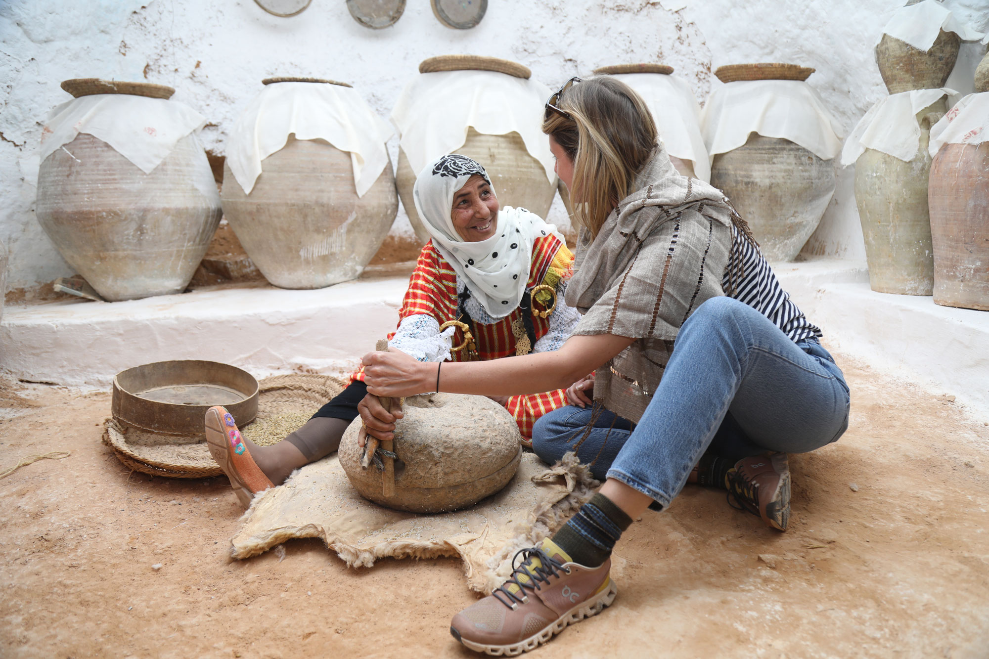 Two women sit on the ground outdoors. On the right, one wears a white head scarf and smiles at a blonde women to her left. Together, they hold a large, round clay mill used to grind couscous. It is placed atop a lambskin rug.