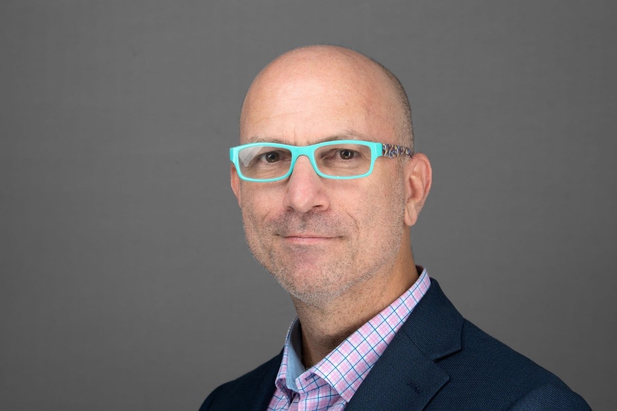 A man in a deep blue dress suit smiles at the camera. He wears bright baby blue reading glasses.