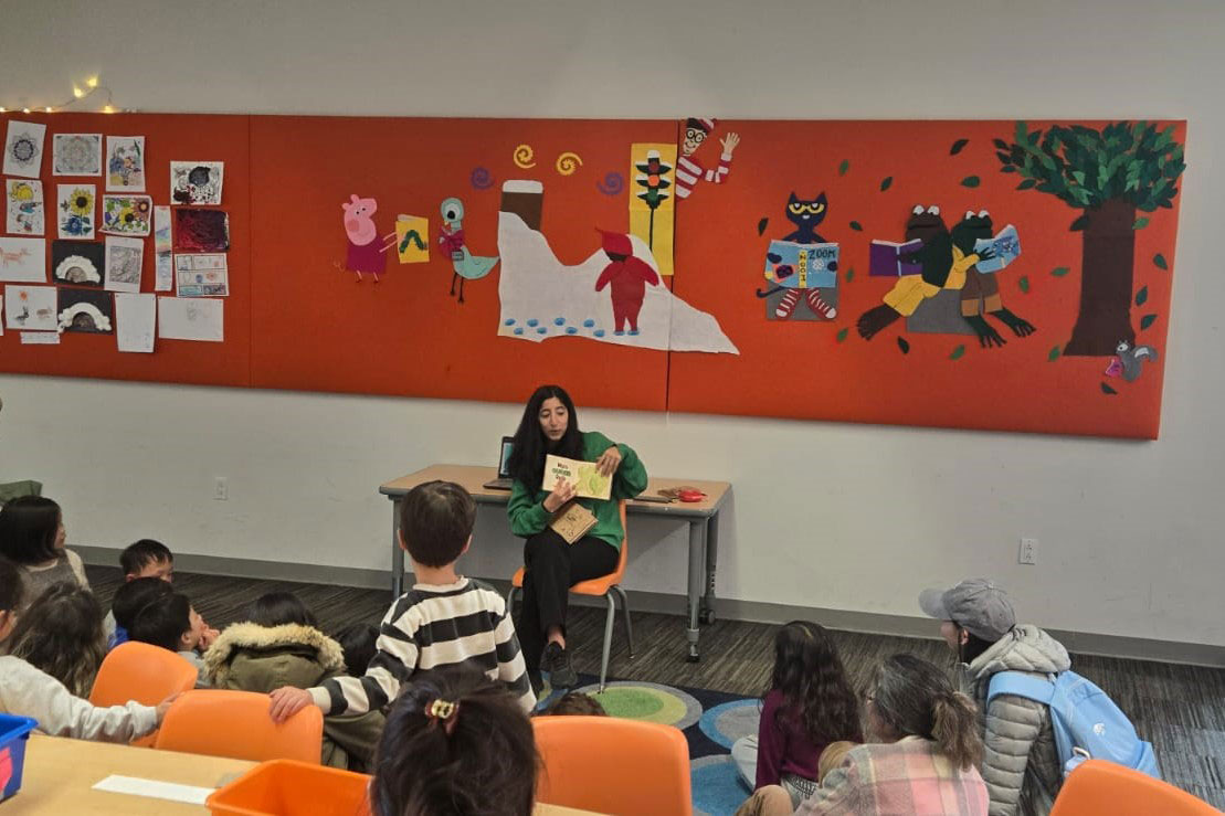 An adult sits in a classroom while reading a book. Children sit on a rug and listen.