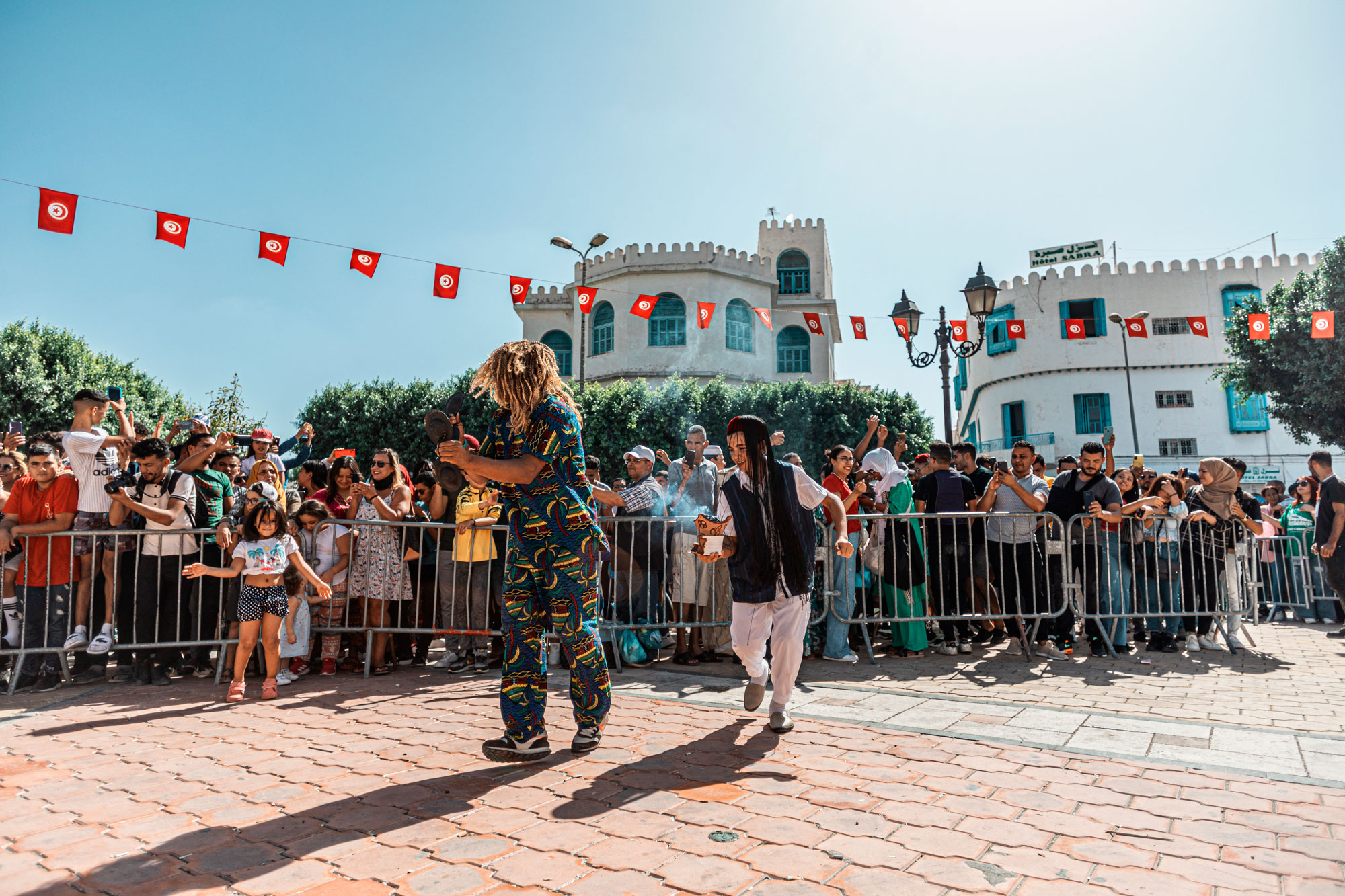 In the mid-day sun, two performers dance in a cobble-stoned town square. Above them hang small Tunisian flags and behind them people of all ages clap and watch, smiling.