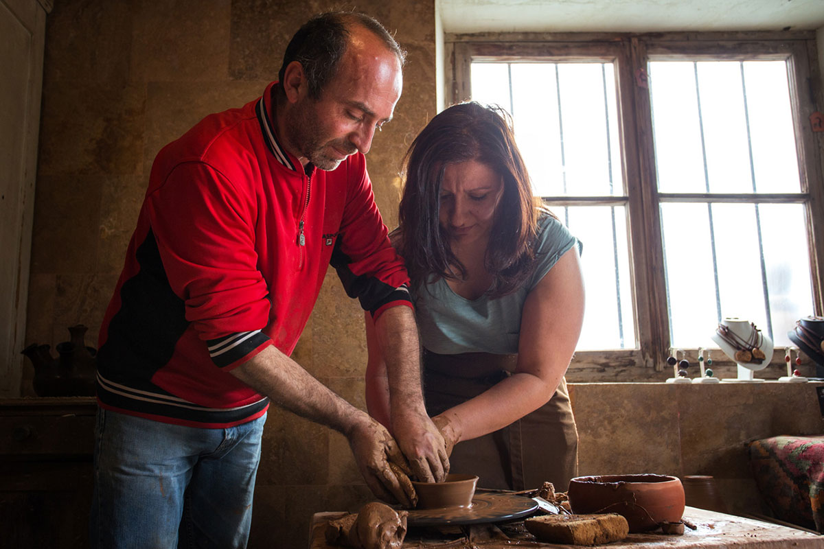 In a rustic room lit by two windows, a man in a red jacket and blue jeans guides the hands of the woman next to him, in a blue blouse and brown apron, both shaping a clay bowl on a spinning pottery wheel. Scattered around them are hunks of clay, finished pottery pieces, and a jewelry display in the windowsill. 