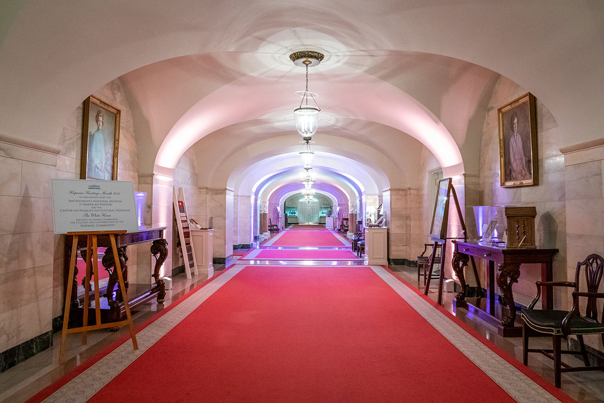 View down a long corridor with white marble walls and red carpet, with display cases and tables on either side. 