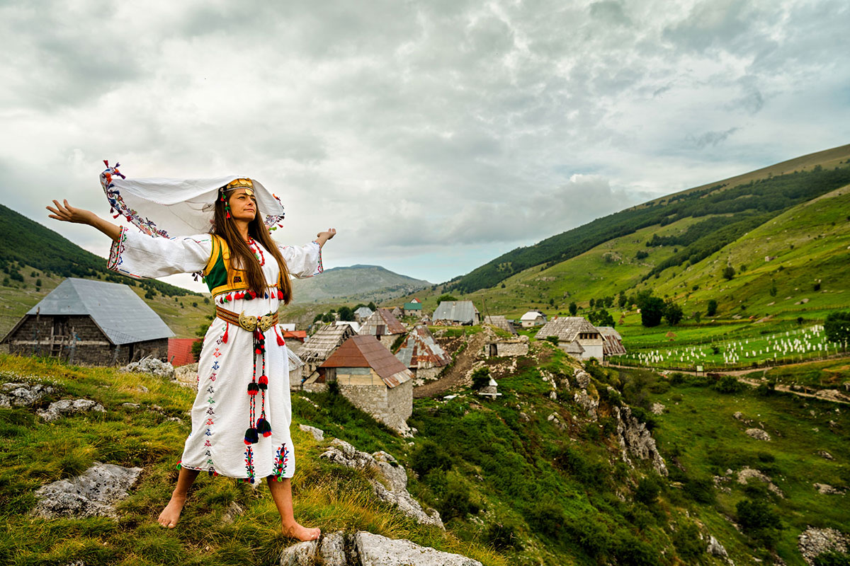 A young woman with long brown hair stands on a scenic village hilltop with her arms outstretched. She wears traditional Bosnian dress which includes a white veil, dress, and green vest, all with colorful embroidery.